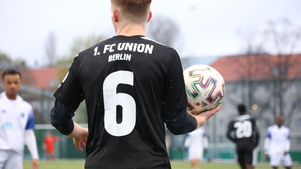 Ein Fußballspieler mit der Nummer 6 in einem schwarzen Trikot hält einen Ball in der Hand, im Hintergrund sind weitere Spieler zu sehen.