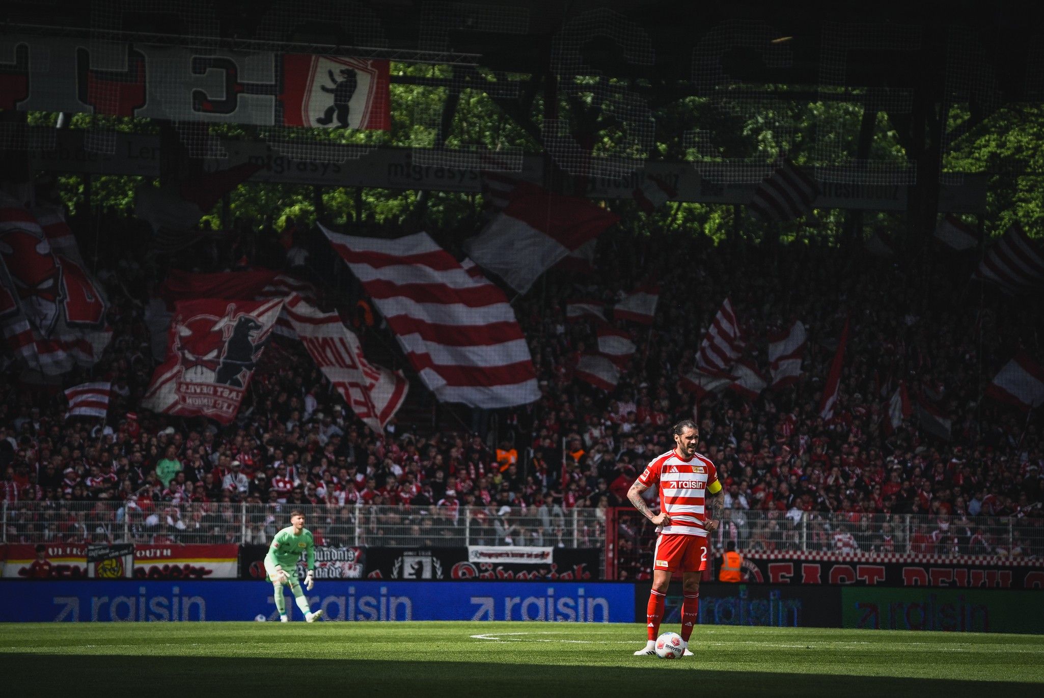 Spielszene im Stadion mit einem Spieler in rot-weißer Uniform, einer Torwartposition und Fans mit Fahnen im Hintergrund.