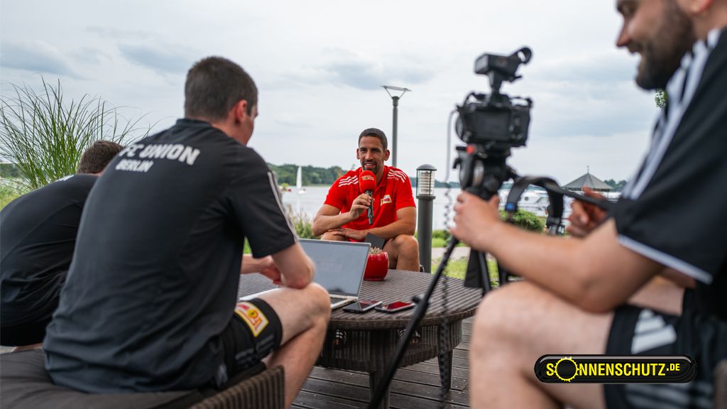 A man in a red jersey sits at a table while being interviewed by several people with a camera and a laptop.