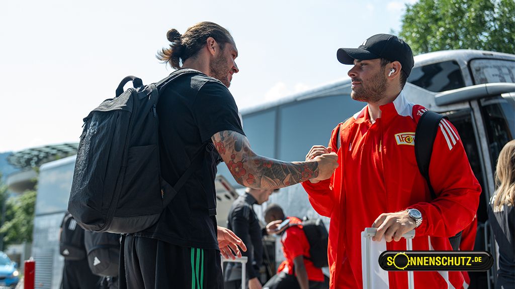 Two men in sportswear are facing each other and greeting each other, with a bus and a sunny environment in the background.