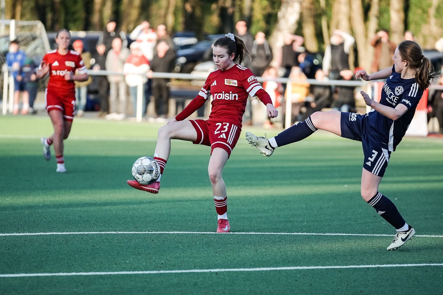 Junge Fußballspielerin in Rot kontrolliert den Ball, während eine Gegnerin in Blau einen Schuss ausführt. Zuschauer im Hintergrund.