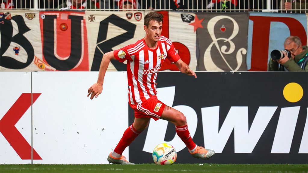 Fußballspieler im roten und weißen Trikot dribbelt den Ball auf dem Spielfeld, im Hintergrund Fankurve mit Banner.