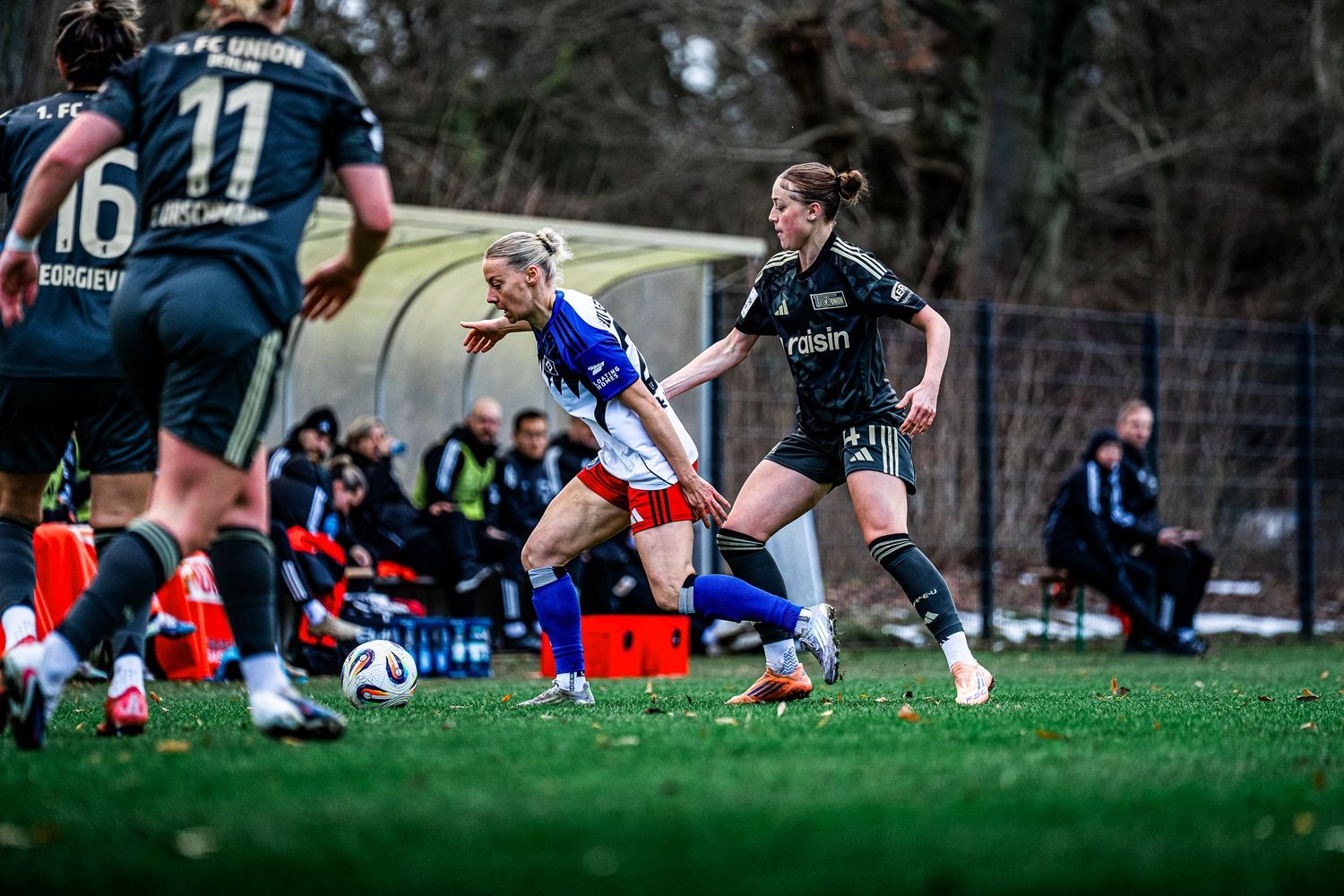 Spielszene im Frauenfußball: Eine Spielerin dribbelt den Ball, während eine Gegnerin sie verfolgt. Zuschauer und Trainer im Hintergrund.