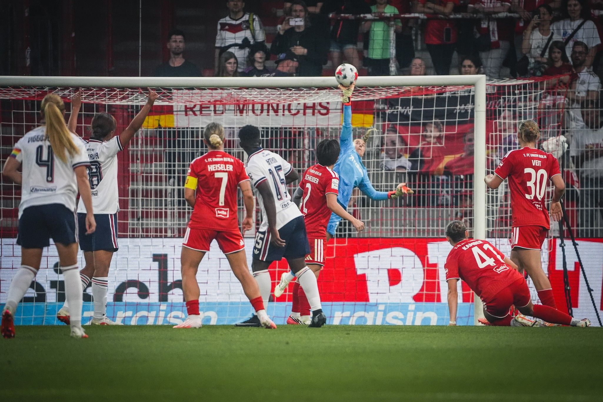 Spielszene im Fußball mit Torwart, der einen Ball abwehrt, und mehreren Spielern, die um das Tor herum positioniert sind.