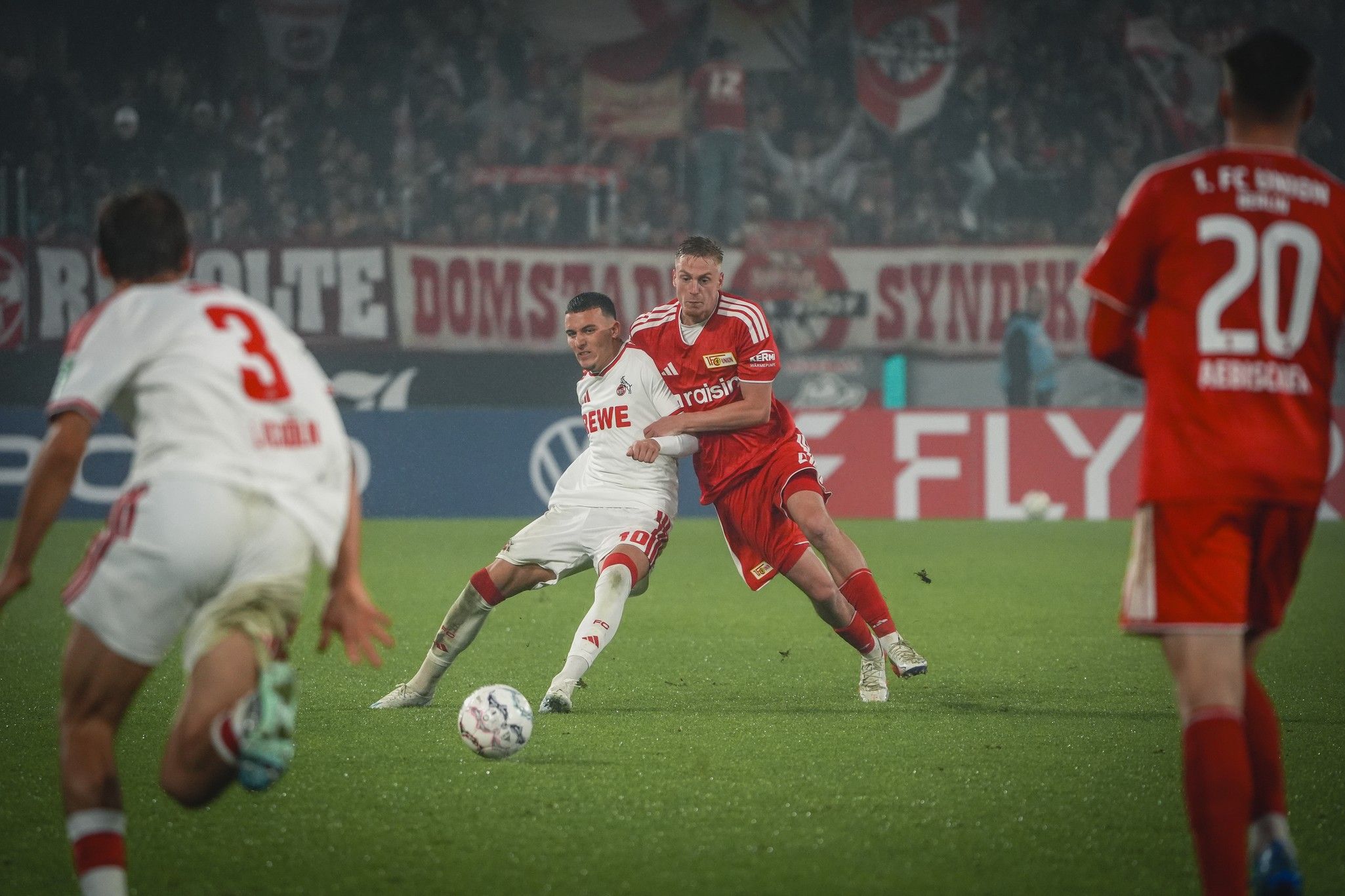 Ein Spieler in rotem Trikot kämpft um den Ball mit einem Spieler in weißem Trikot, während Zuschauer im Hintergrund zu sehen sind.