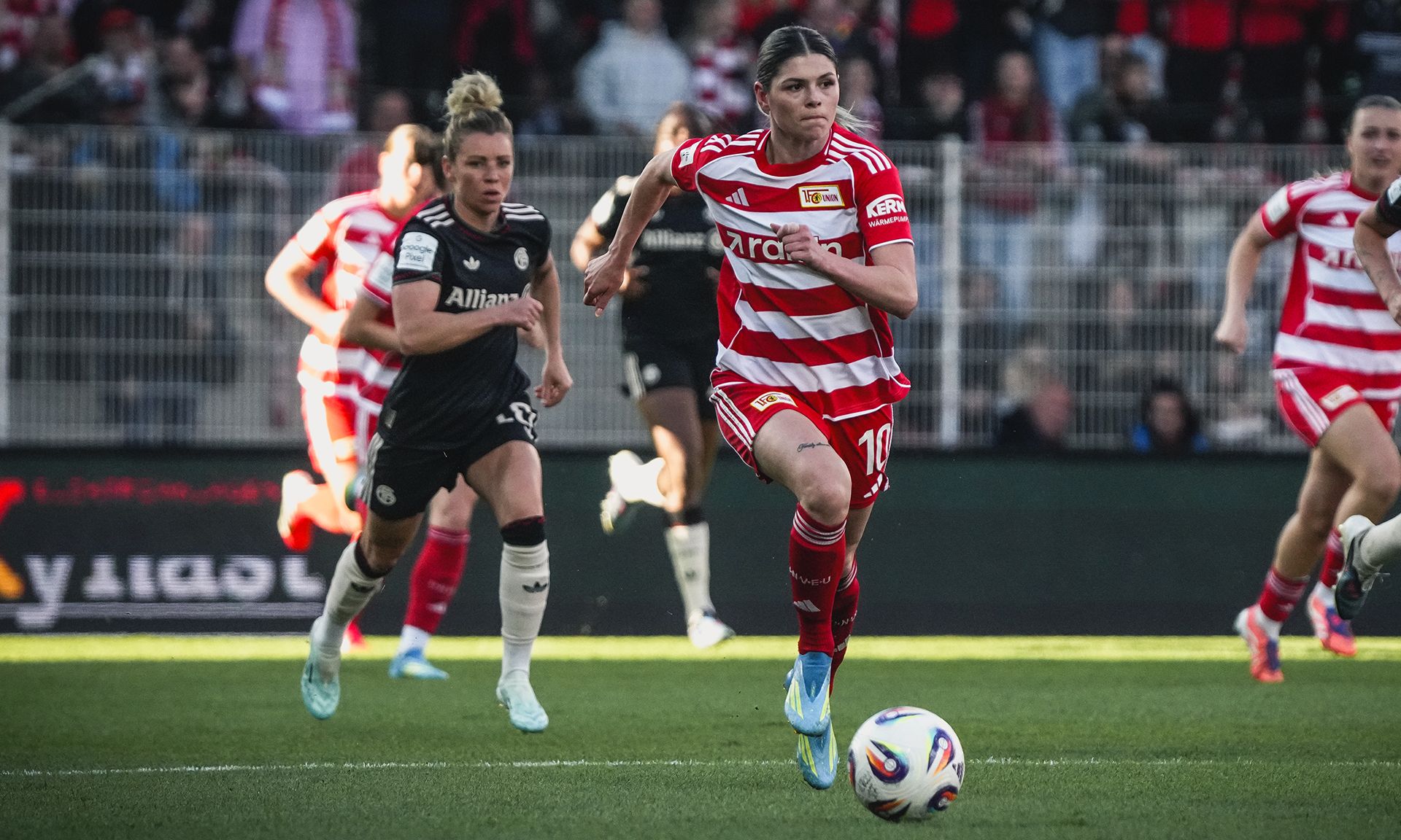 Spielszene im Frauenfußball: Eine Spielerin in gestreifter roter und weißer Dress sprintet mit dem Ball, während eine gegnerische Spielerin folgt.