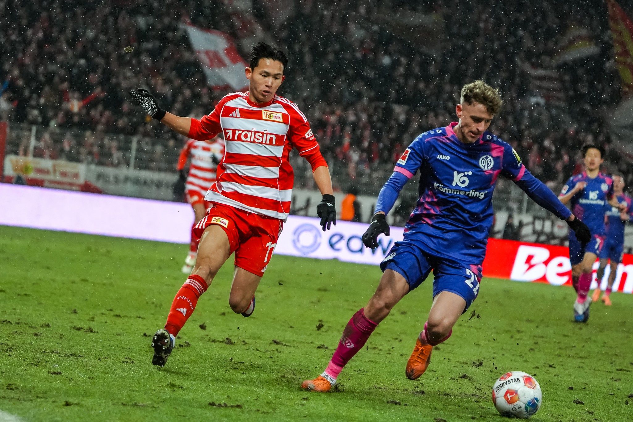 Zwei Fußballspieler im Wettkampf um den Ball auf einem nassen Spielfeld während eines Spiels. Fans im Hintergrund.