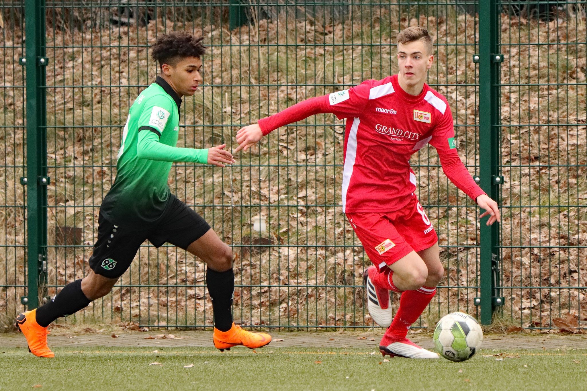 A football match between two players, one in a red jersey and the other in green-black, on a grass field.