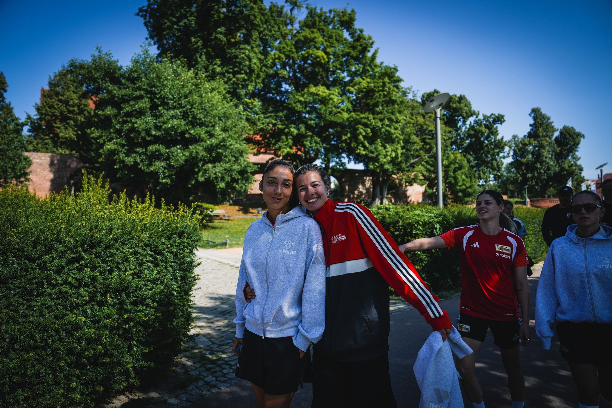 Zwei Personen in Sportkleidung umarmen sich lächelnd, während sie an einem sonnigen Tag in einem Park spazieren.