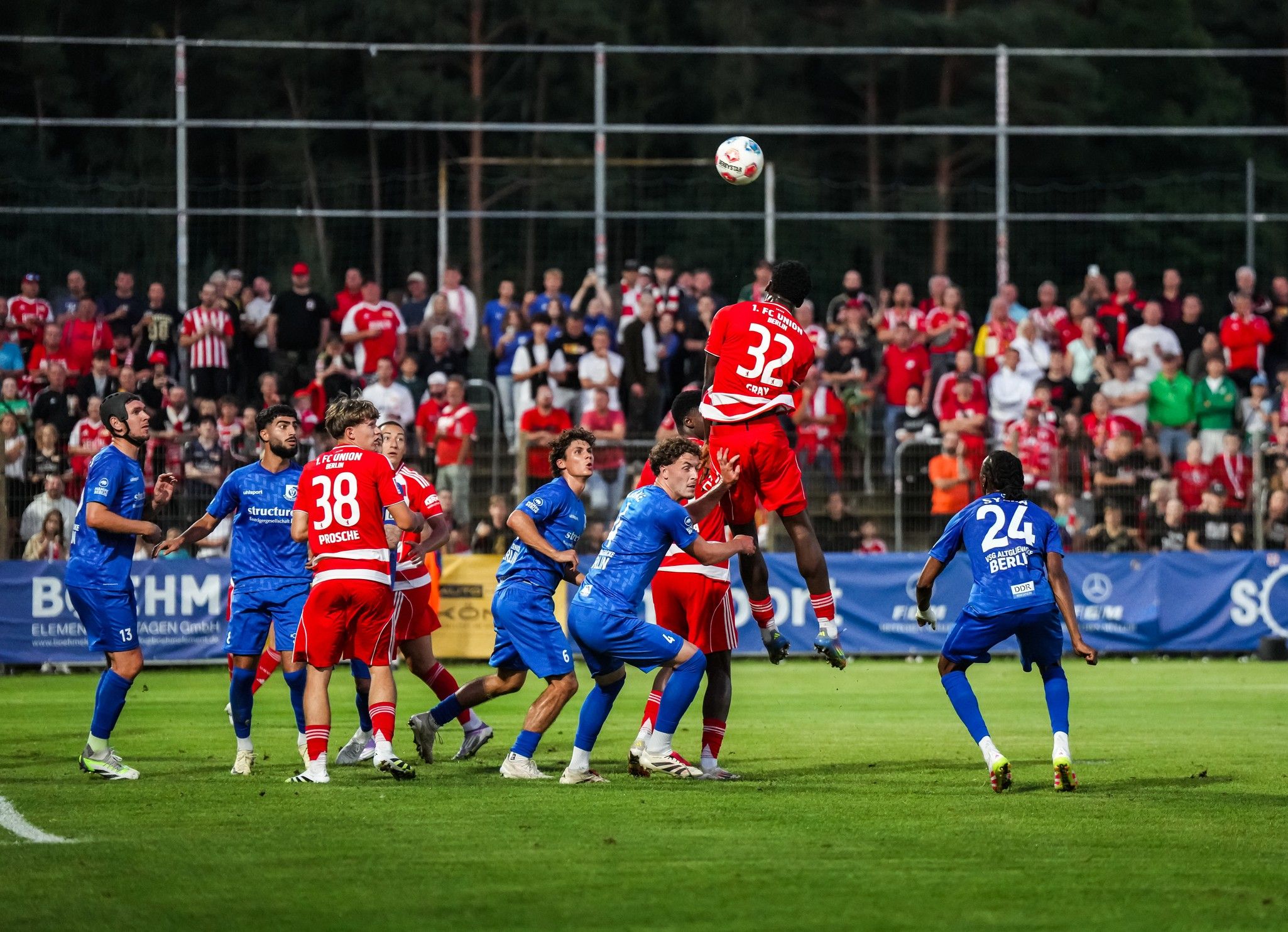 Ein Spieler in roter Kleidung springt hoch, um einen Ball während eines Fußballspiels zu köpfen, während mehrere Spieler in blauer Kleidung versuchen, ihn zu blocken.