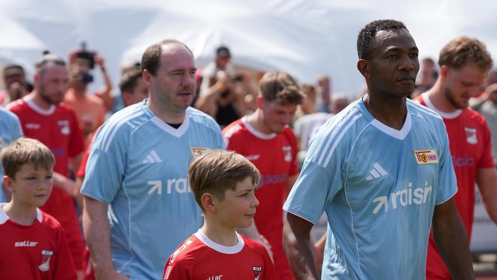 Spieler betreten das Spielfeld, begleitet von Kindern, in Fußballtrikots vor einer jubelnden Menge.