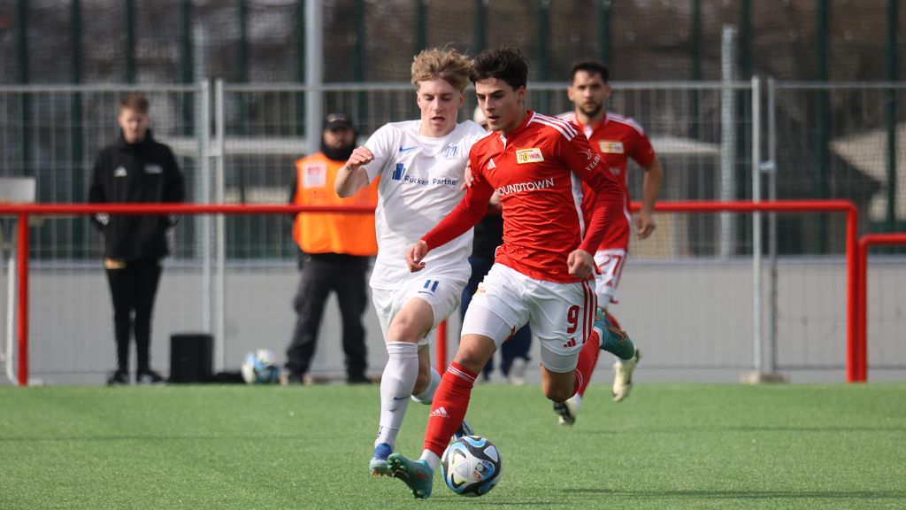 Zwei Fußballspieler kämpfen um den Ball auf einem grünen Kunstrasenfeld. Ein Spieler trägt ein rotes Trikot, der andere ein weißes.