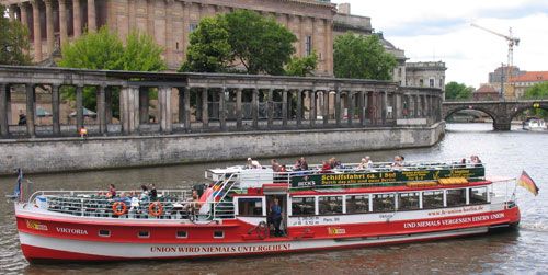 Rotes Ausflugsschiff mit Passagieren auf einem Fluss, im Hintergrund historische Gebäude und Bäume.