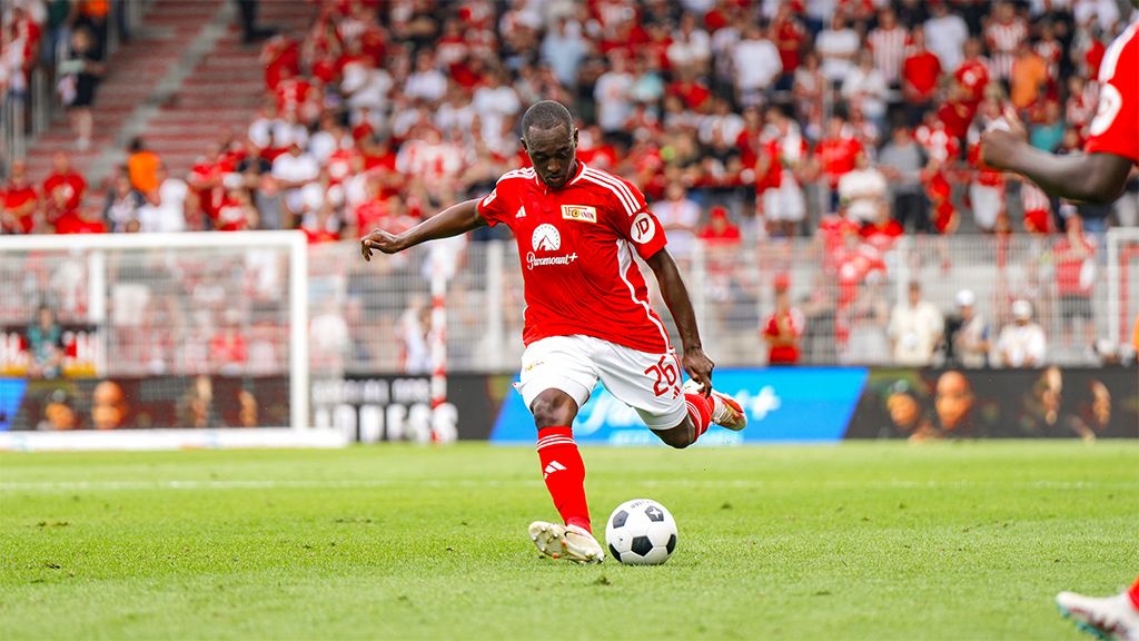 Fußballspieler mit der Nummer 25 in rotem Trikot schießt einen Ball während eines Spiels vor jubelndem Publikum.