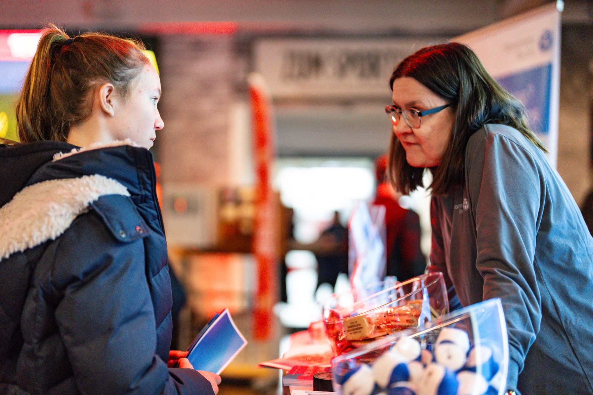 Zwei Personen stehen an einem Messestand, eine junge Frau in einem blauen Mantel spricht mit einer Frau am Stand.