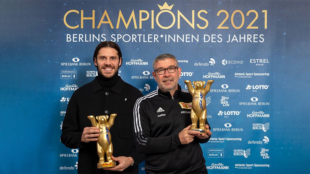Zwei Männer halten goldene Trophäen vor einem Hintergrund mit dem Schriftzug "Champions 2021".