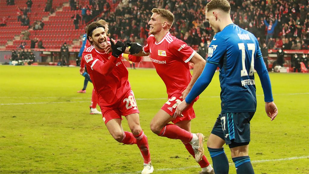 Zwei Spieler in roten Trikots feiern jubilierend auf dem Fußballfeld, während ein Spieler in Blau im Hintergrund steht.