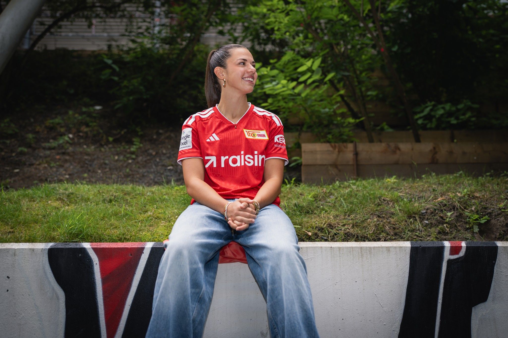 Eine Person sitzt lächelnd auf einer Bank, trägt ein rotes Fußballtrikot und Jeans, umgeben von grüner Vegetation.