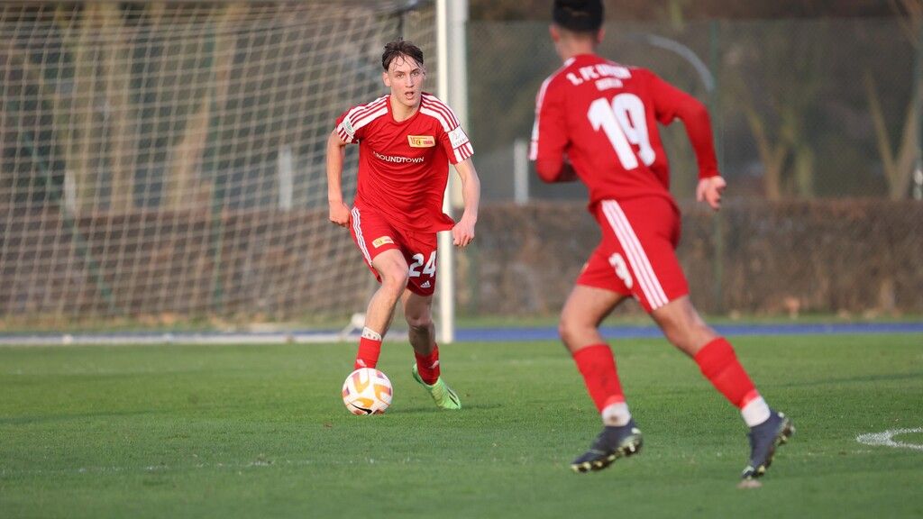 Zwei Fußballspieler in roten Trikots dribbeln den Ball auf einem grünen Spielfeld. Im Hintergrund steht ein Tor.