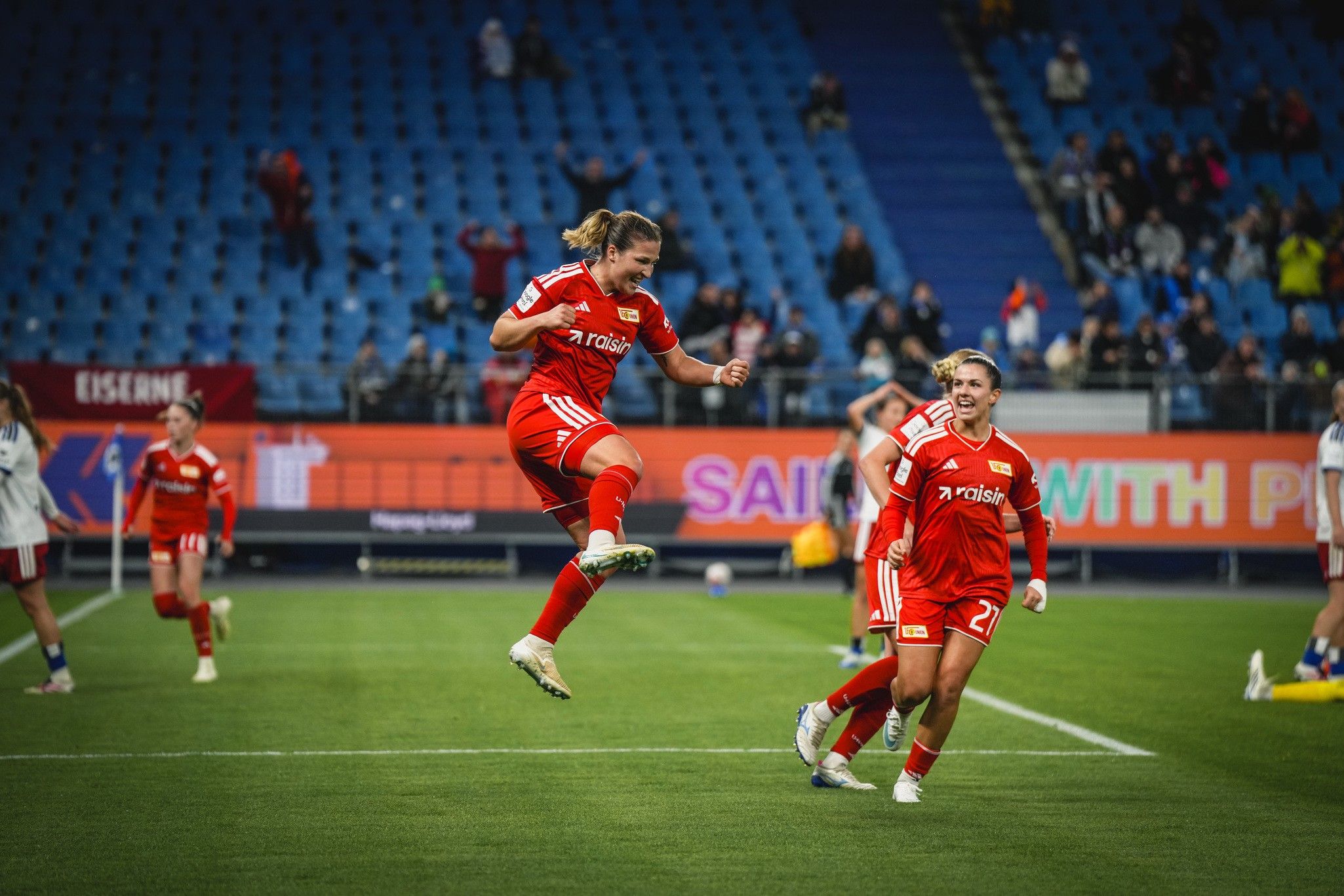Eine Spielerin in rotem Trikot springt jubelnd auf dem Fußballfeld, während ihre Teamkollegin zusieht.