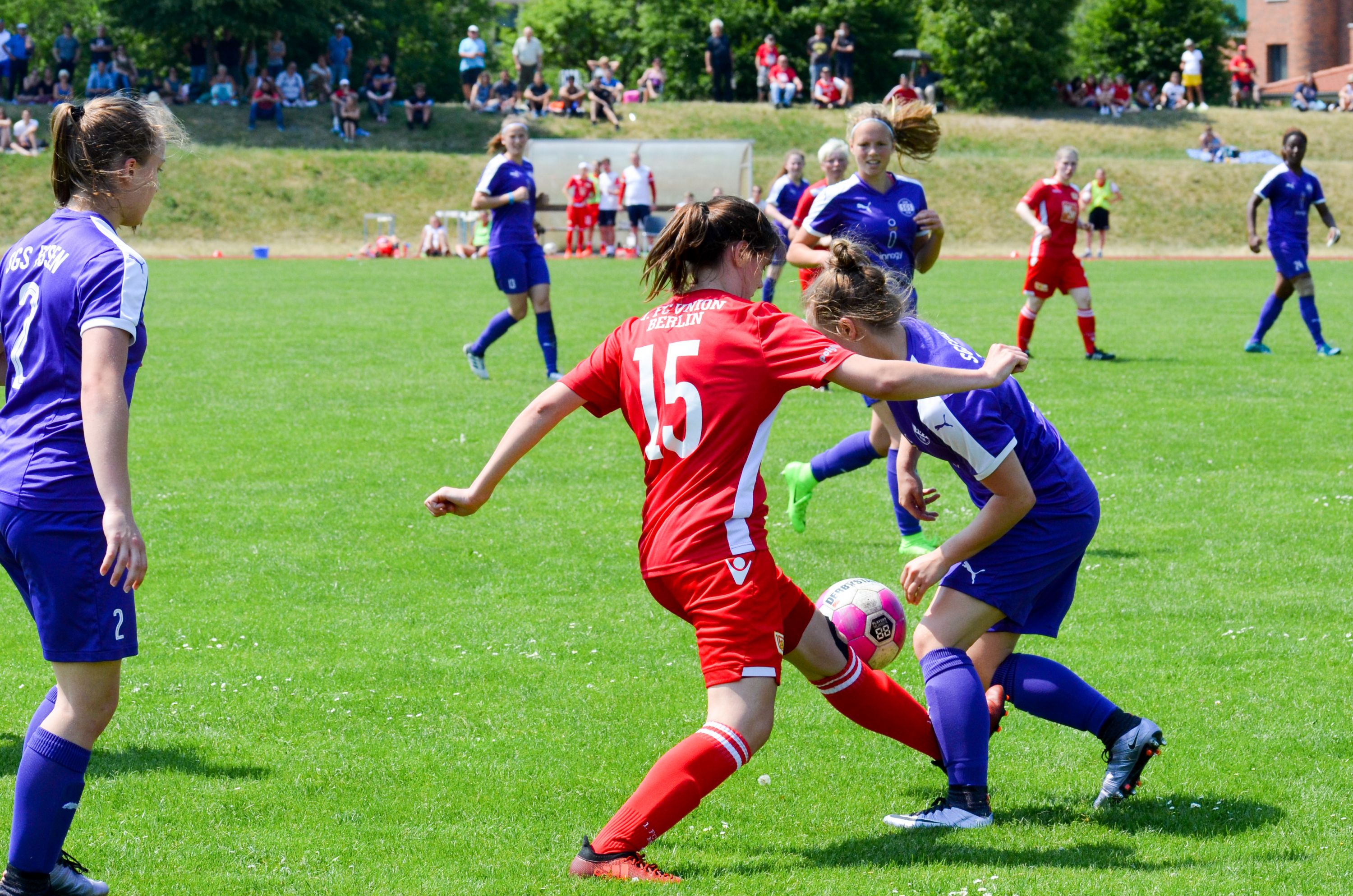 Zwei junge Frauen spielen Fußball auf einem grünen Platz, während Zuschauer im Hintergrund zuschauen. Eine Spielerin trägt ein rotes, die andere ein lila Trikot.