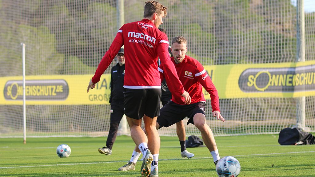 Zwei Fußballspieler in Trainingsanzügen üben Passspiel auf einem Rasenplatz, im Hintergrund ist ein Trainer zu sehen.