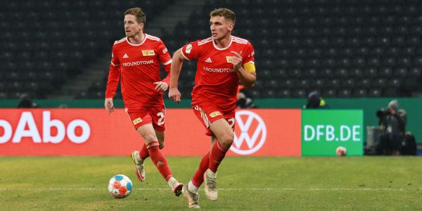 Zwei Fußballspieler in roten Trikots von Union Berlin laufen über das Spielfeld, während ein Ball vor ihnen rollt.