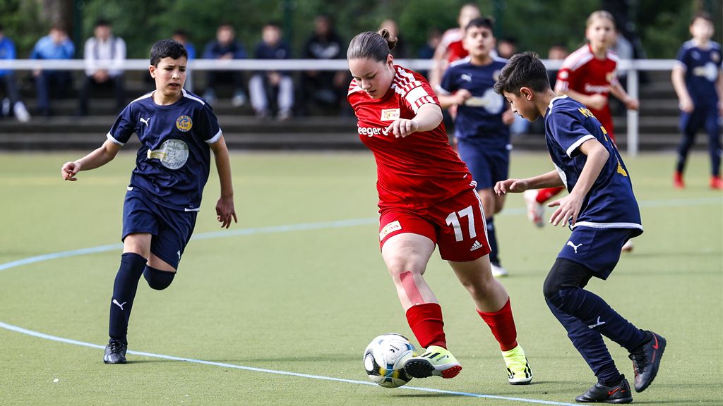 Ein Jugendfußballspiel auf einem Kunstrasenplatz, zwei Spieler im blauen Trikot versuchen, den Ball von einer Spielerin im roten Trikot zu erobern.