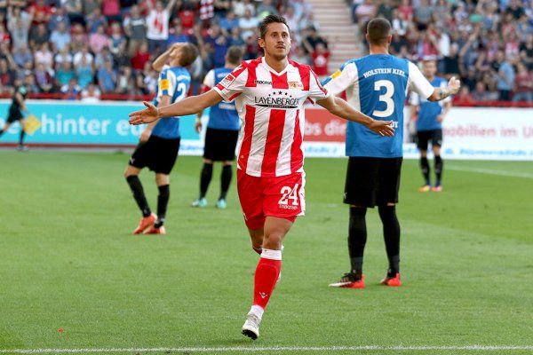 A football player in a red and white jersey celebrates a goal while teammates and opponents react in the background.