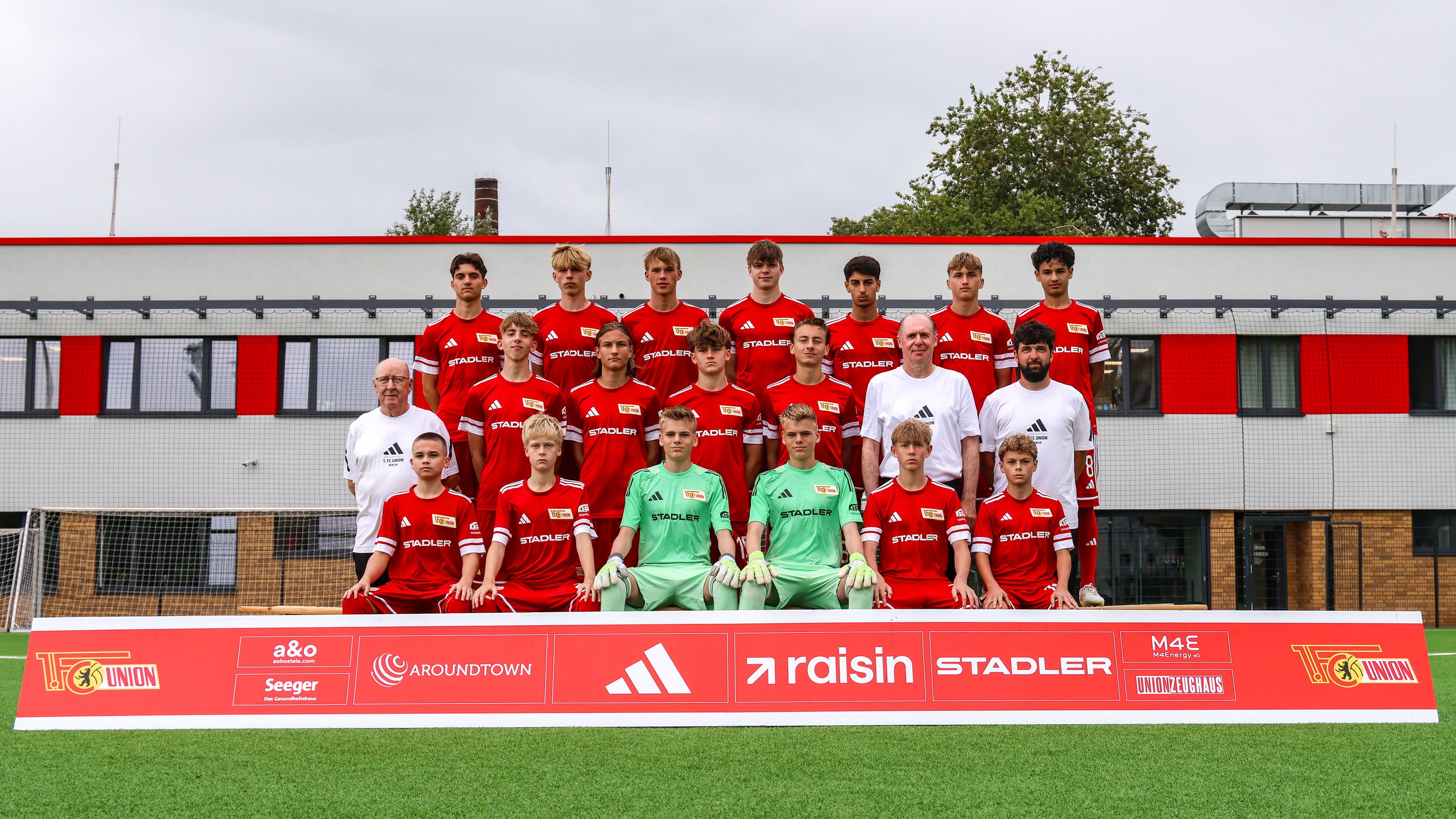 Eine Gruppenaufnahme einer Fußballmannschaft in roten Trikots, stehend und sitzend, vor einem Trainingsgelände.