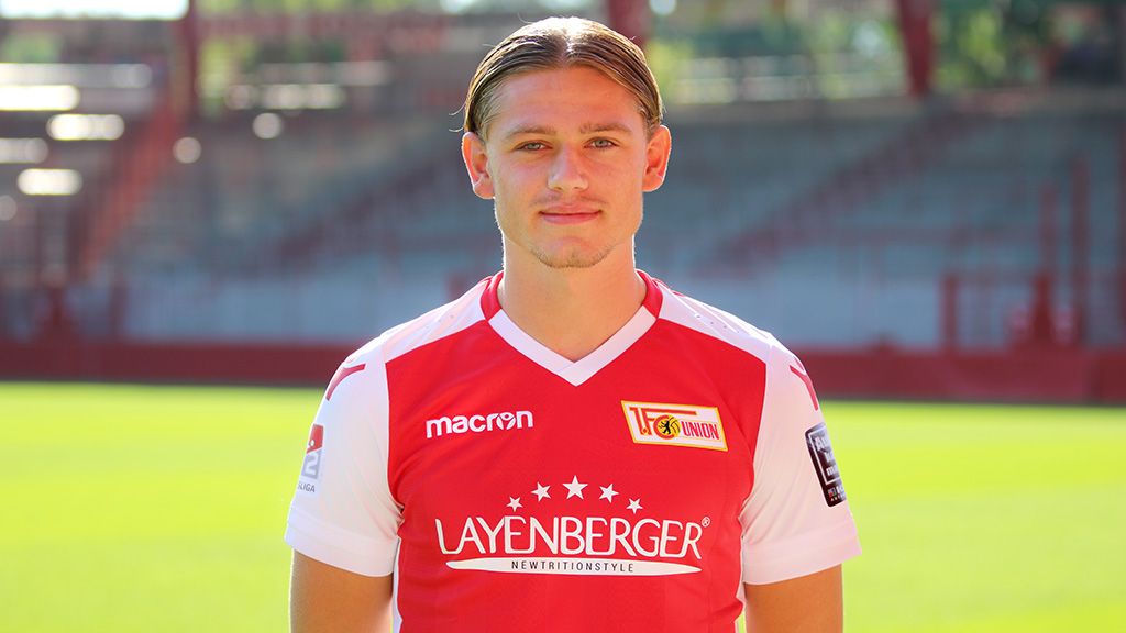 A young football player in a red jersey stands before a stadium backdrop, smiling at the camera.