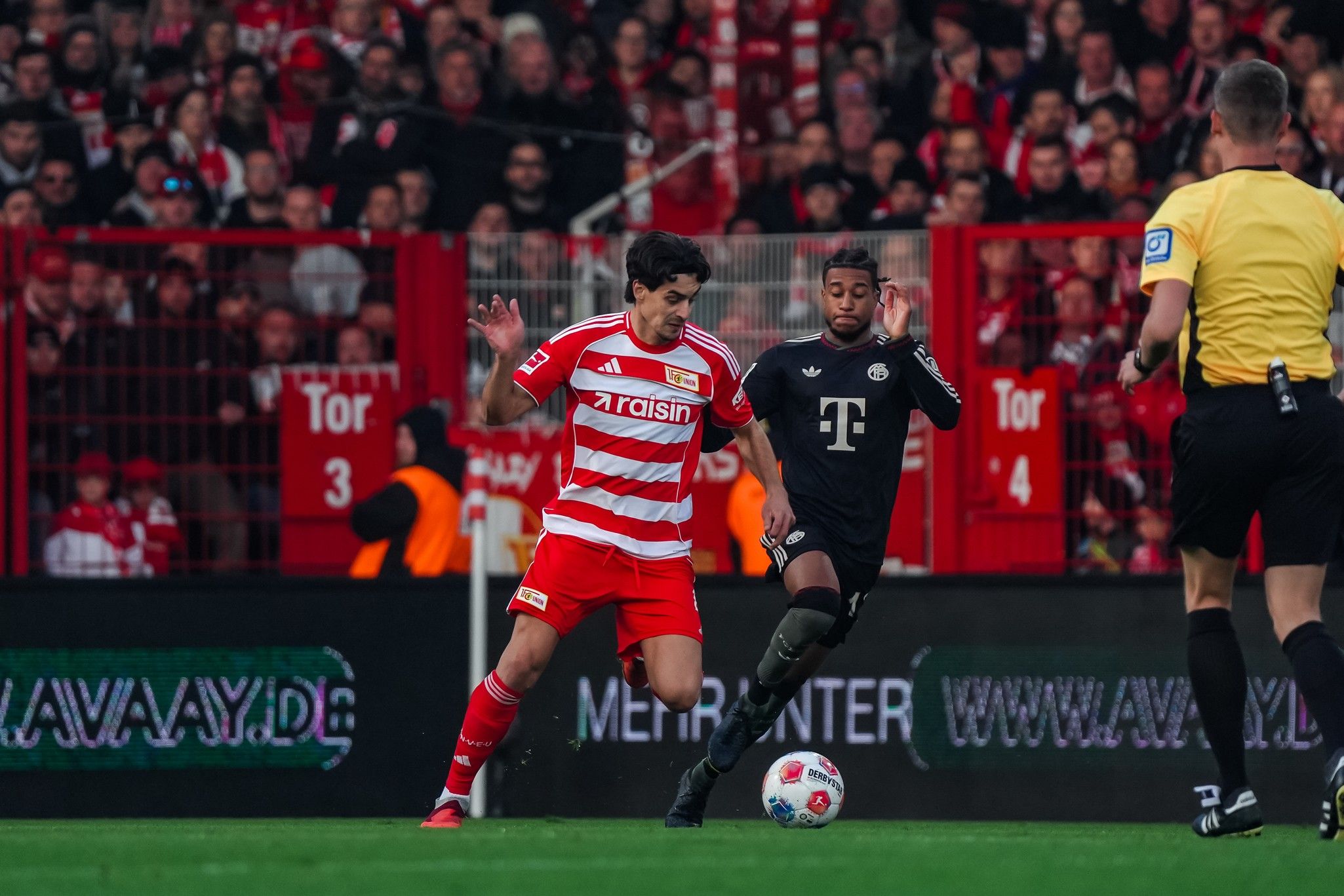 Ein Fußballspiel zwischen zwei Mannschaften, ein Spieler in rot-weiß gestreiftem Trikot dribbelt, der andere in schwarz daneben.