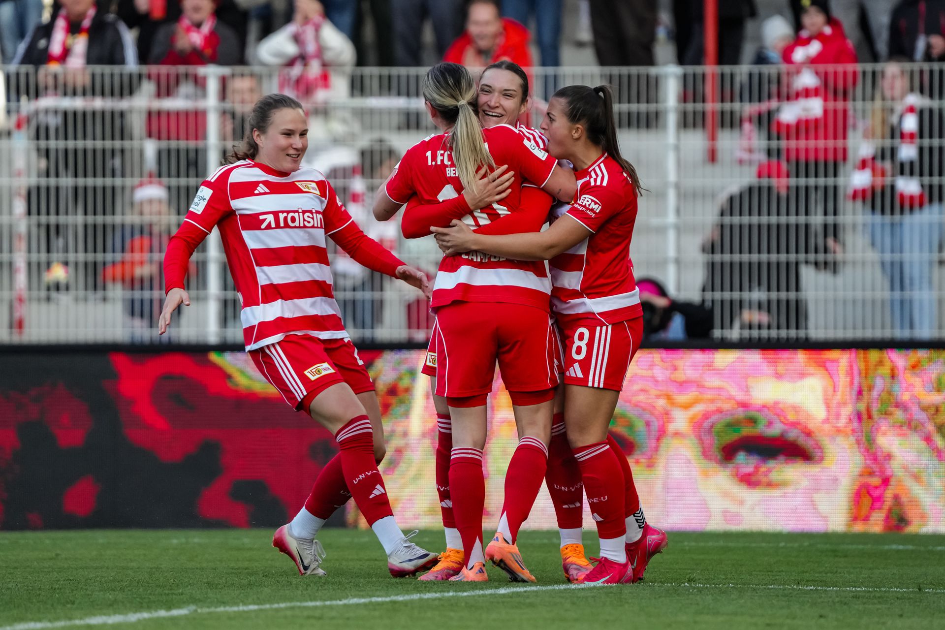 Vier Fußballspielerinnen in roten Trikots feiern einen Torerfolg auf dem Spielfeld. Zuschauer im Hintergrund.