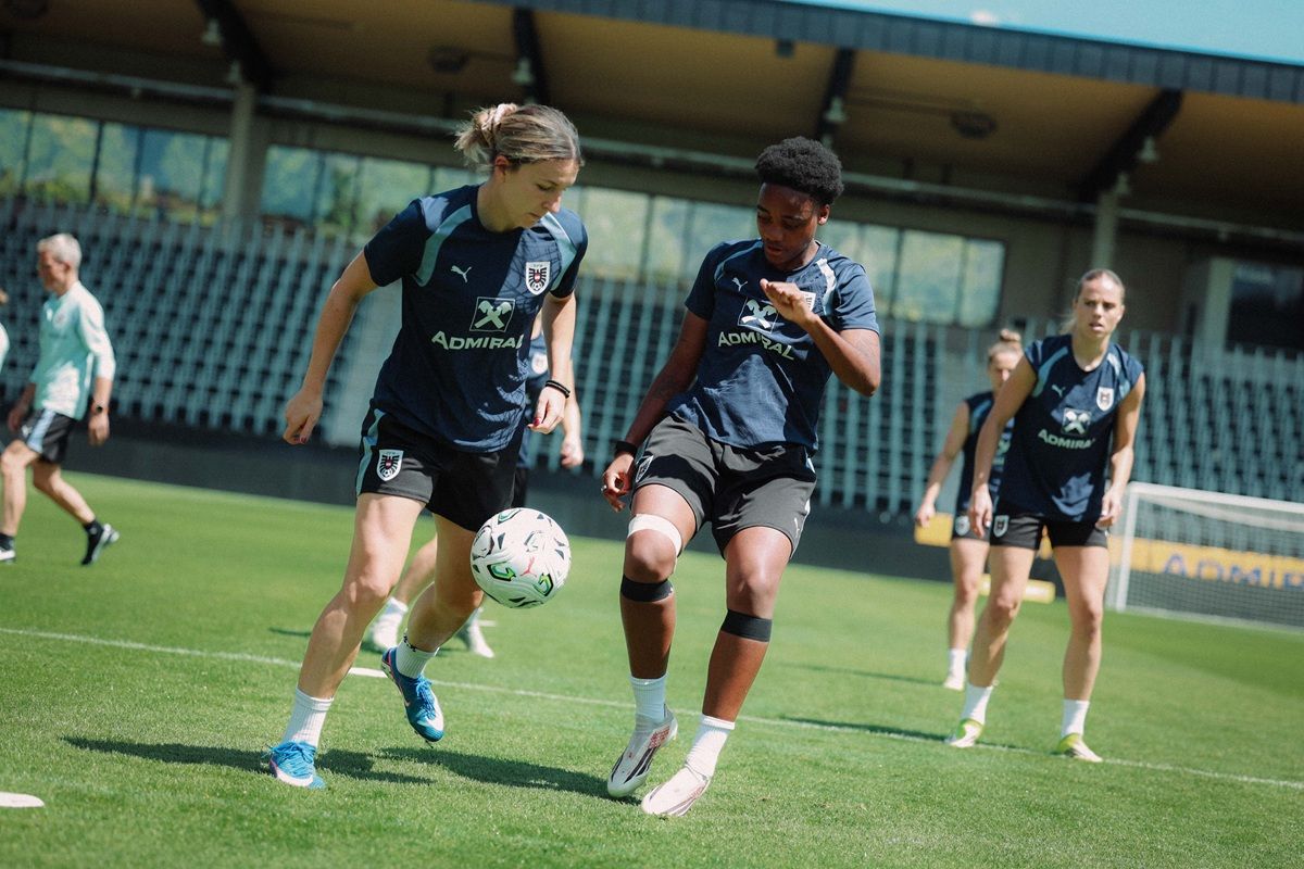 Zwei Frauen trainieren auf einem Fußballfeld, während eine dritte im Hintergrund zusieht.