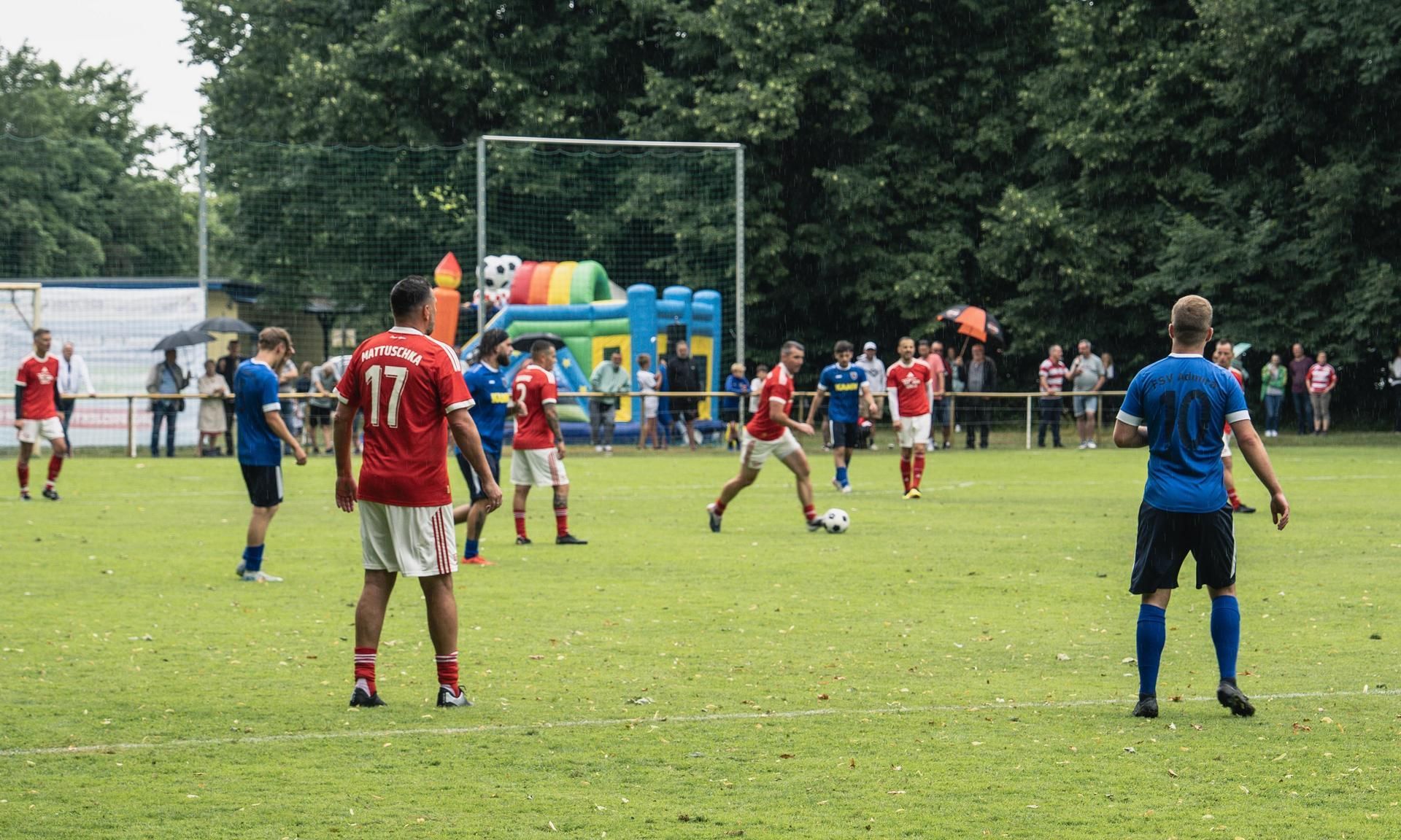 Spielszene auf einem Fußballfeld mit Spielern in roten und blauen Trikots, im Hintergrund ein Spielplatz.