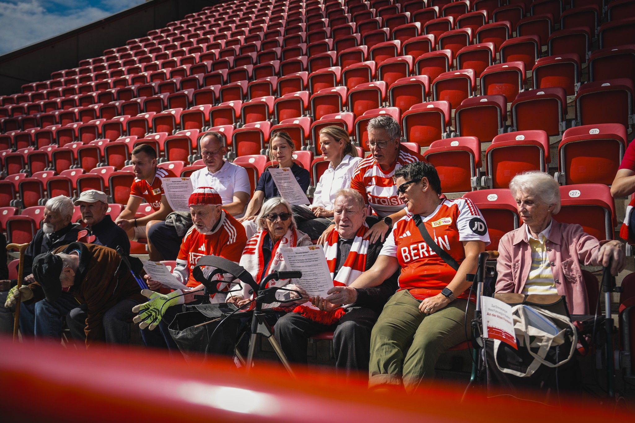 Eine Gruppe von Menschen in roten Trikots sitzt auf roten Stadionbänken und hält Liederblätter in den Händen.