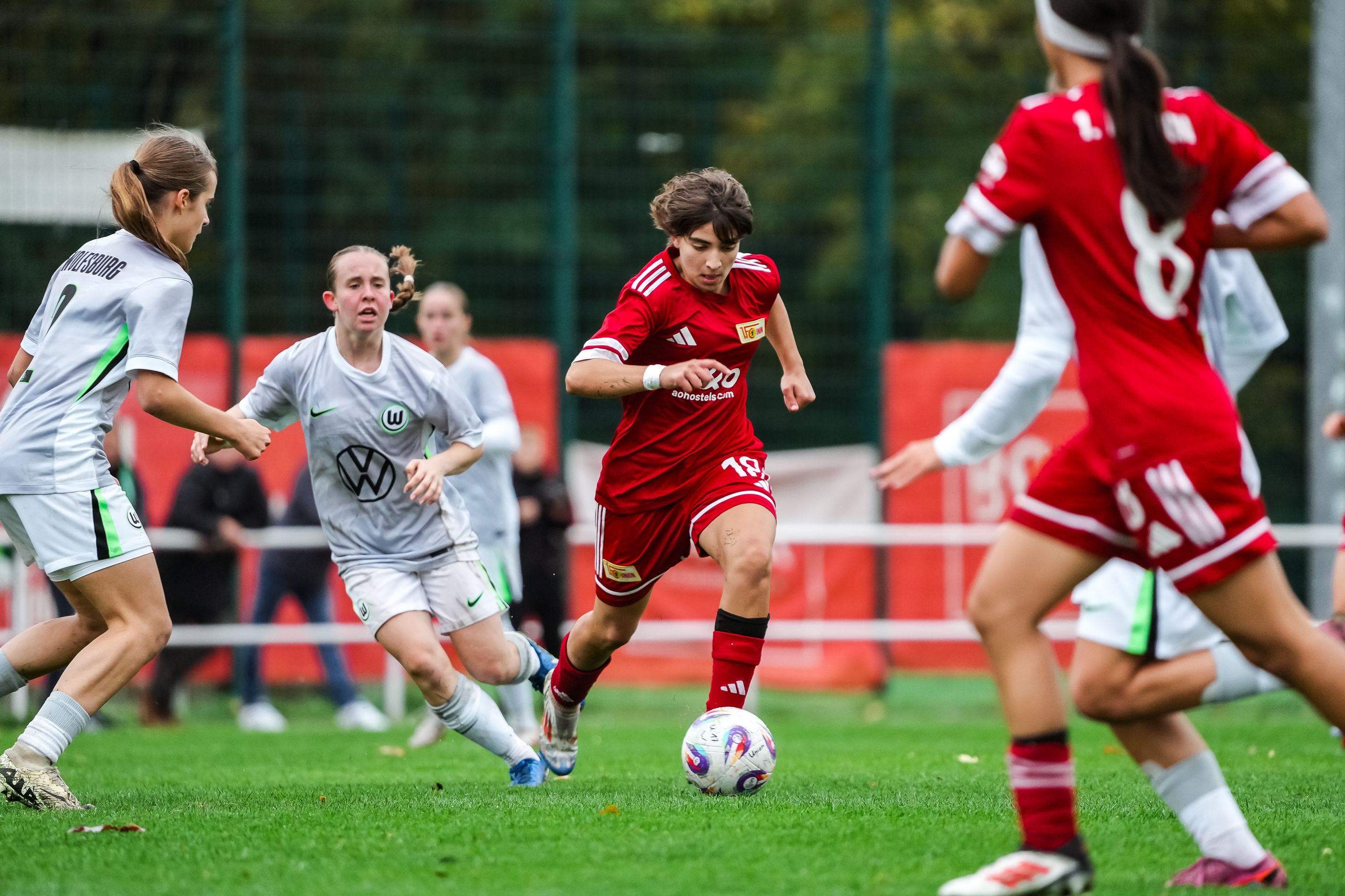 Eine Spielerin in rotem Trikot dribbelt mit dem Ball, während zwei Gegnerinnen in weißen Trikots verfolgen. Fußballspiel auf einem Rasenplatz.