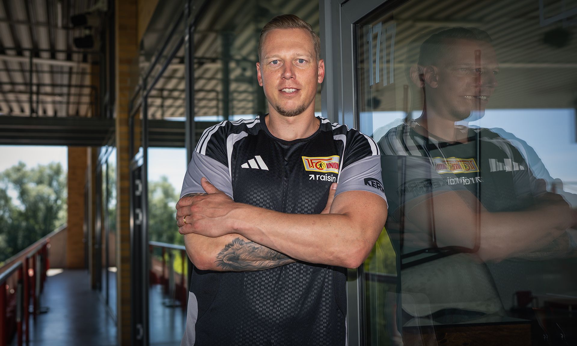 Ein Mann in Sportbekleidung steht vor einem Fenster und lächelt, mit Armen verschränkt. Im Hintergrund ist ein grünes Landschaftsbild sichtbar.
