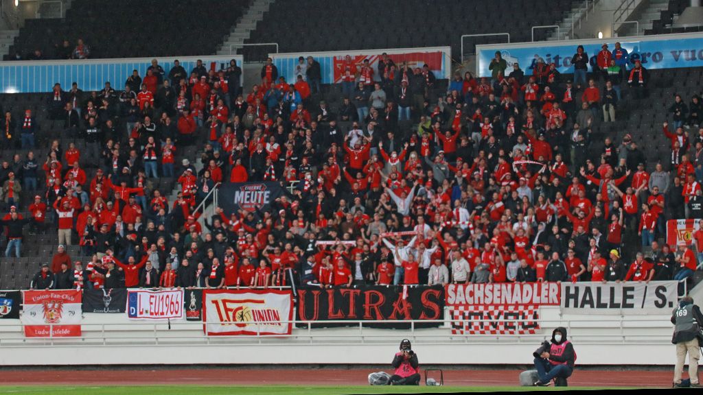 Group of cheering fans in red jerseys holding flags and banners in a stadium during a game.