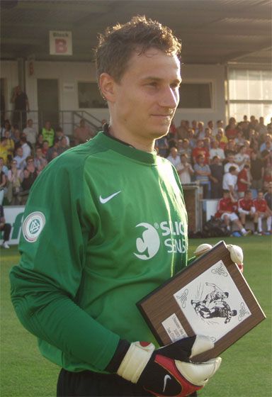 Ein Fußballtorwart in grüner Trikots mit Handschuhen hält eine Auszeichnung in der Hand, während eine Menschenmenge im Hintergrund steht.