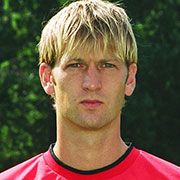 Ein junger Mann mit blonden Haaren trägt ein rotes Sporttrikot und schaut ernst in die Kamera, von einem grünen Hintergrund umgeben.