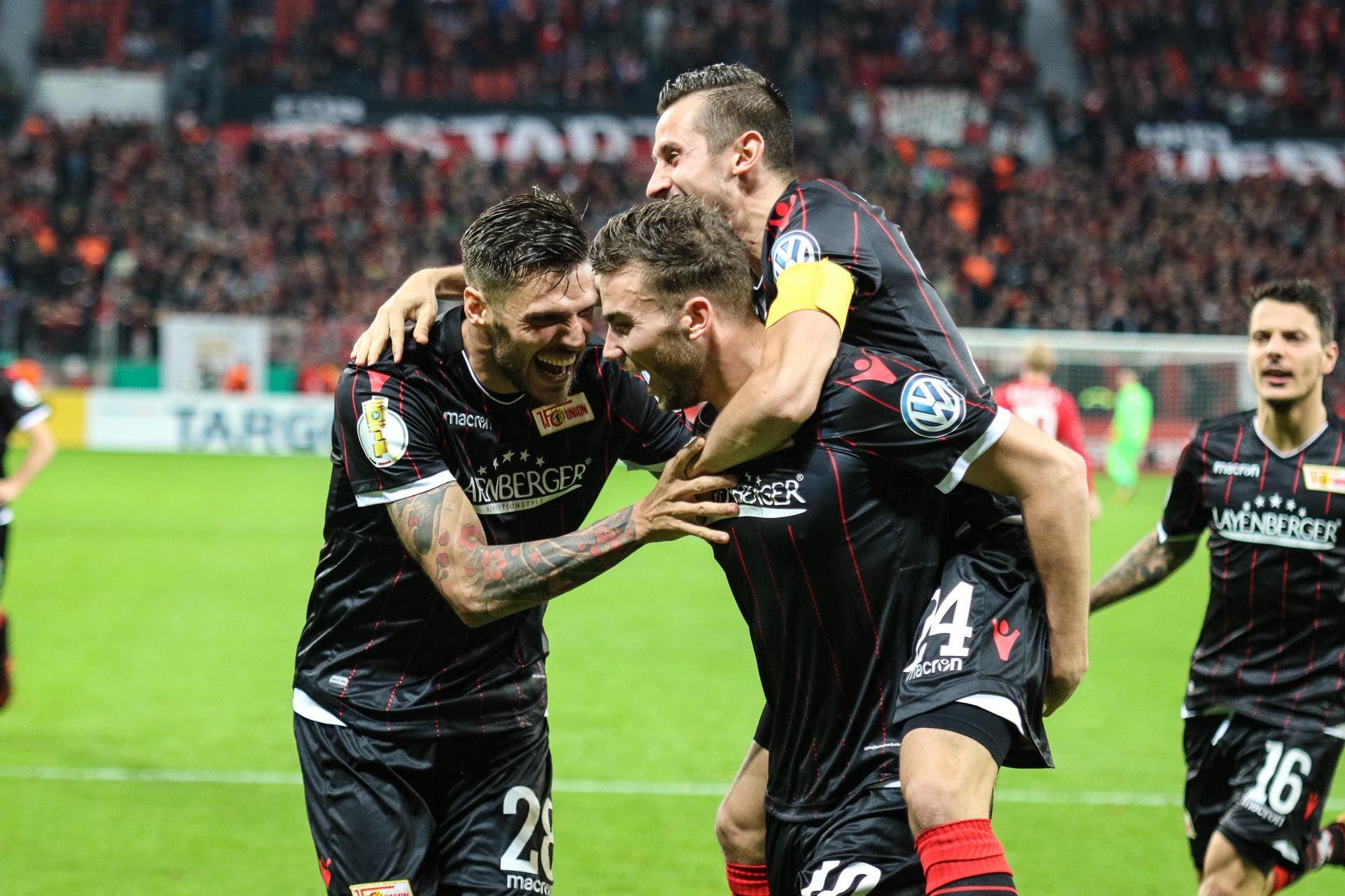 Drei jubelnde Fußballspieler in schwarz-roten Trikots feiern ein Tor vor einer jubelnden Menge im Stadion.