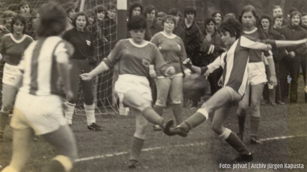 Eine historische Szene eines Frauenfußballspiels mit Spielerinnen in blauen und roten Trikots, umgeben von Zuschauern.