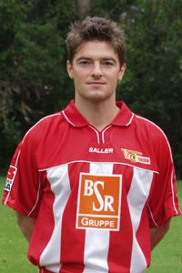 Ein junger Mann in einem rot-weißen Fußballtrikot steht lächelnd vor einem grünen Hintergrund.