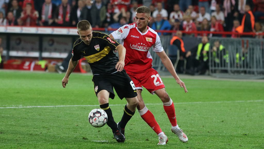 Zwei Fußballspieler in Aktion, einer mit schwarzem Trikot und der andere mit weiß-rotem Trikot, kämpfen um den Ball auf dem Spielfeld.