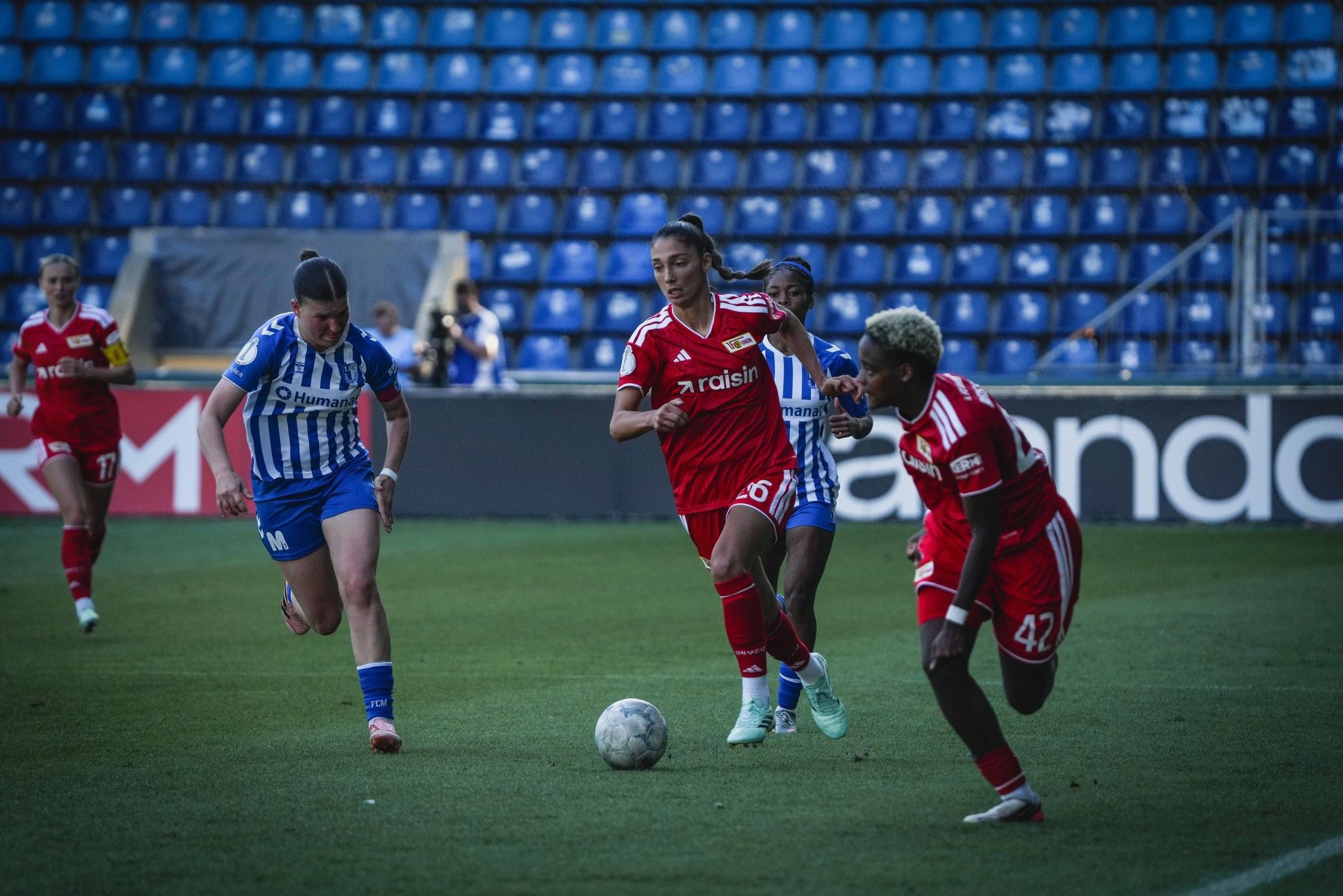 Zwei Fußballspielerinnen in roten Trikots dribbeln den Ball, während eine Spielerin in blauem Trikot verfolgt. Stadionhintergrund.