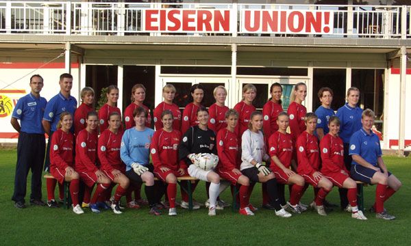 Grupp portrait der Frauenfußballmannschaft von Union Berlin in roten Trikots, mit Trainer und Betreuern im Hintergrund.