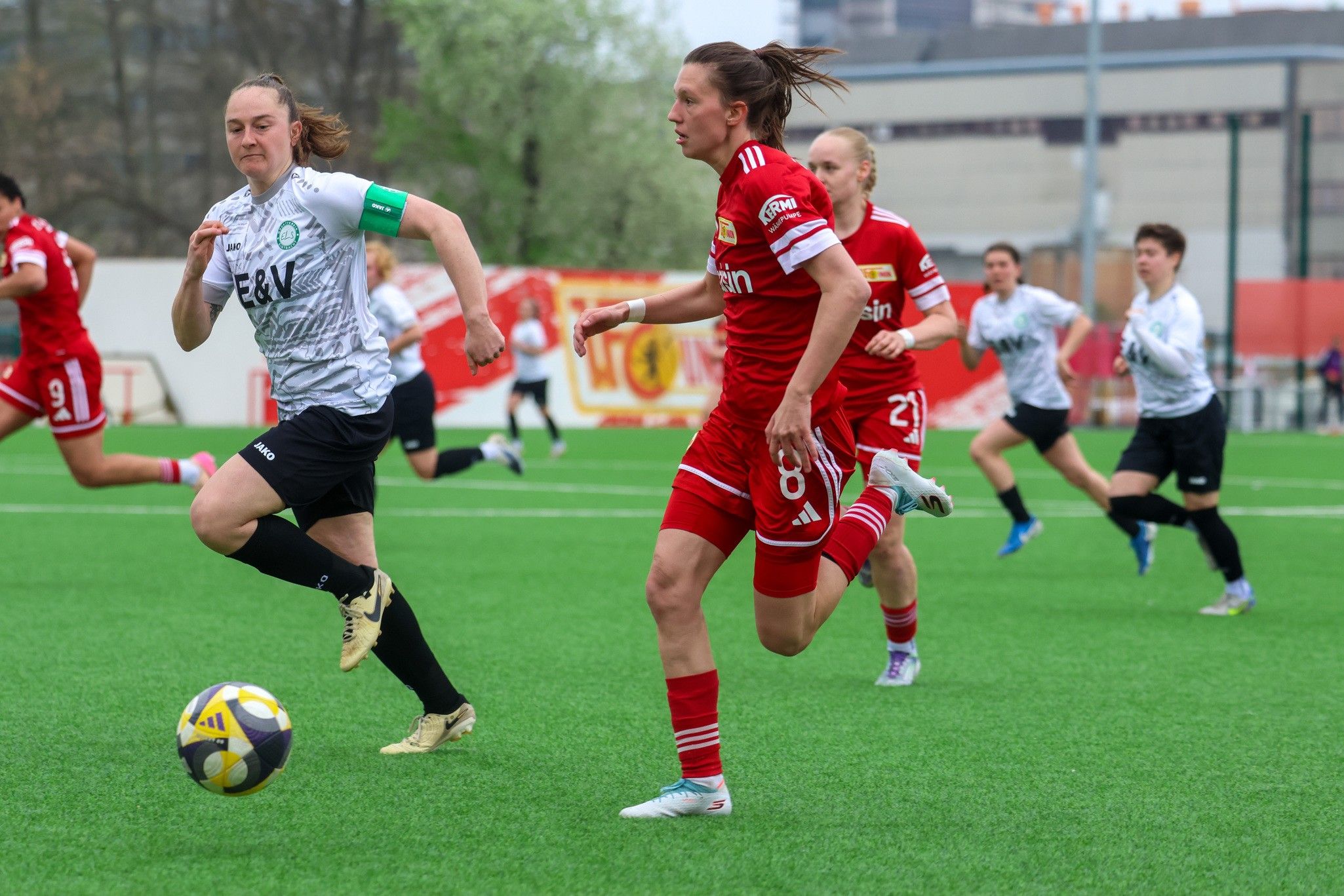 Zwei Fußballspielerinnen sprinten auf einem Kunstrasenplatz, während weitere Spielerinnen im Hintergrund stehen.
