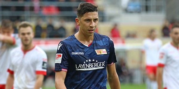 Ein Fußballspieler in blauer Mannschaftskleidung läuft über das Spielfeld, umgeben von anderen Spielern in weißen und roten Trikots.