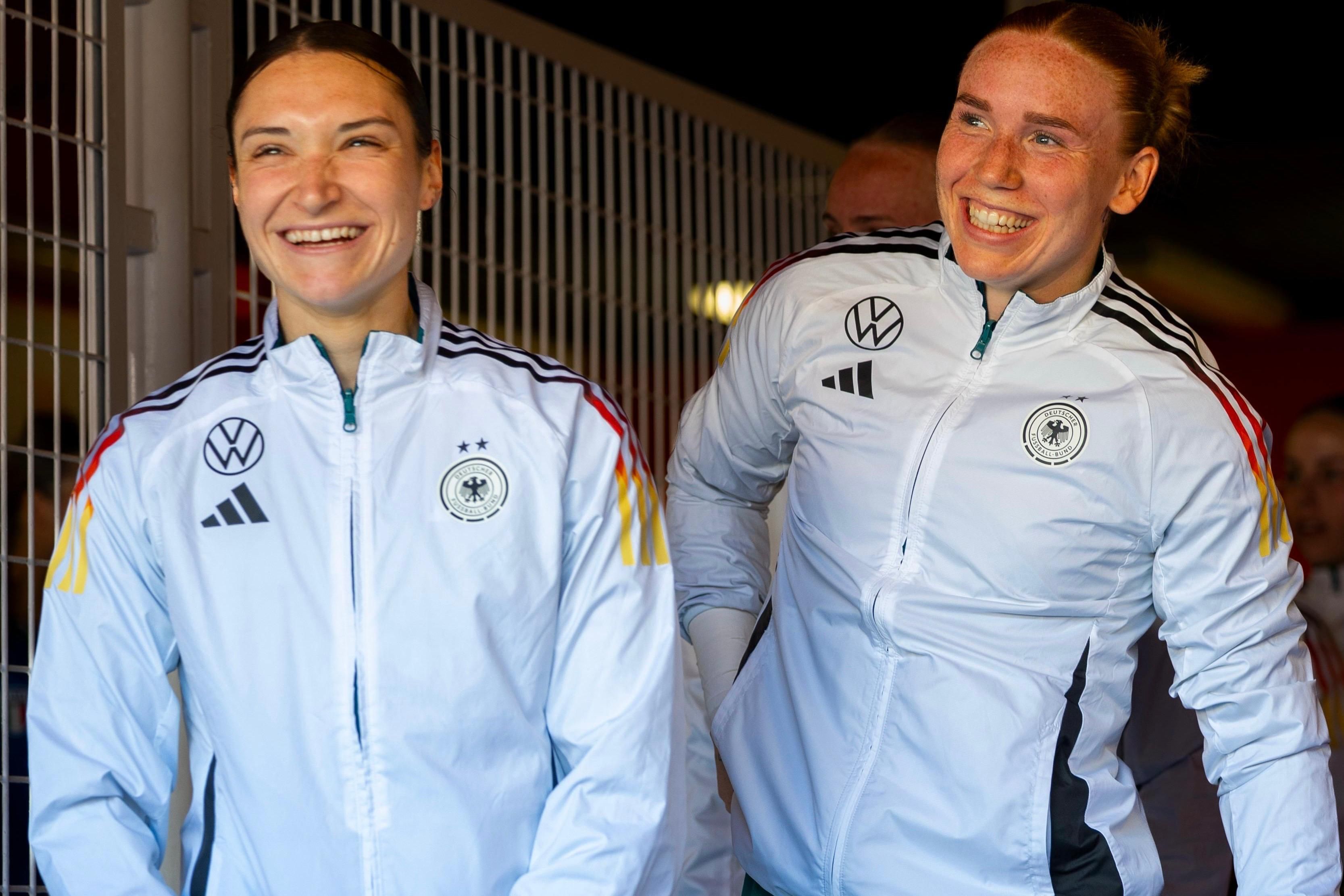 Zwei lächelnde Frauen in weißen Sportjacken mit schwarz-rot-goldenen Elementen betreten einen Sportbereich.